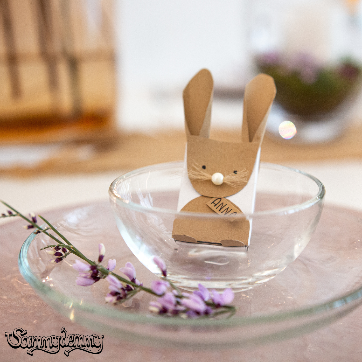 Ein niedlicher Hase aus Kraftpapier sitzt in einer transparenten Glasschale und dient als Namensschild mit der Aufschrift „Anna“ – eine liebevolle Osterdeko.