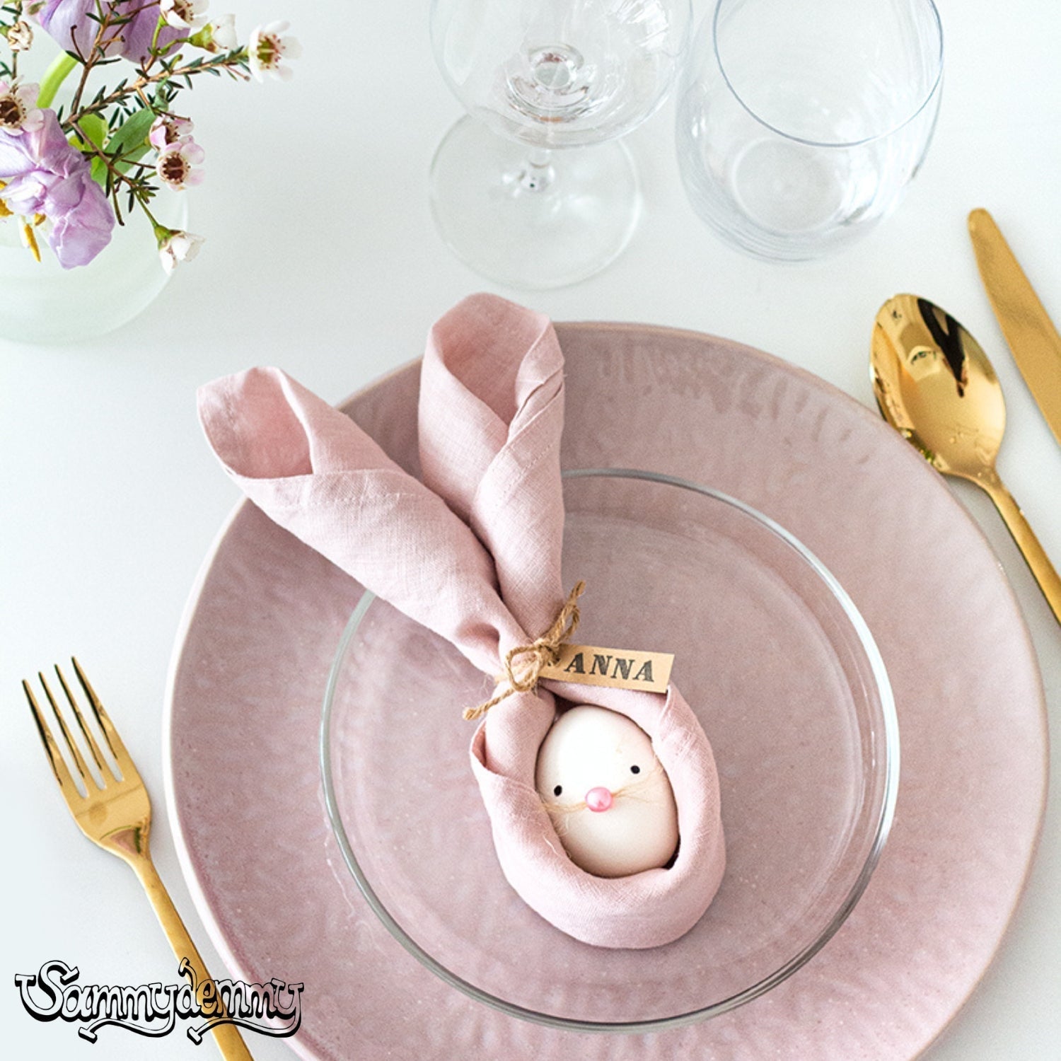 Ein festlich gedeckter Ostertisch mit einem zartrosa Teller-Set, goldfarbenem Besteck und einer liebevoll gestalteten Serviette in Hasenform. In der Serviette ist ein bemaltes Ei mit Hasengesicht eingebunden, versehen mit einem Namensschild „Anna“