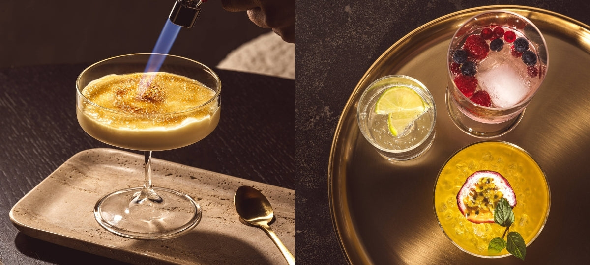 Mehrere stilvoll inszenierte Cocktails in eleganten Gläsern auf dunklem Tisch. Ein Drink wird mit einem Bunsenbrenner karamellisiert, andere sind mit Zitrusfrüchten, Beeren oder Passionsfrucht dekoriert.