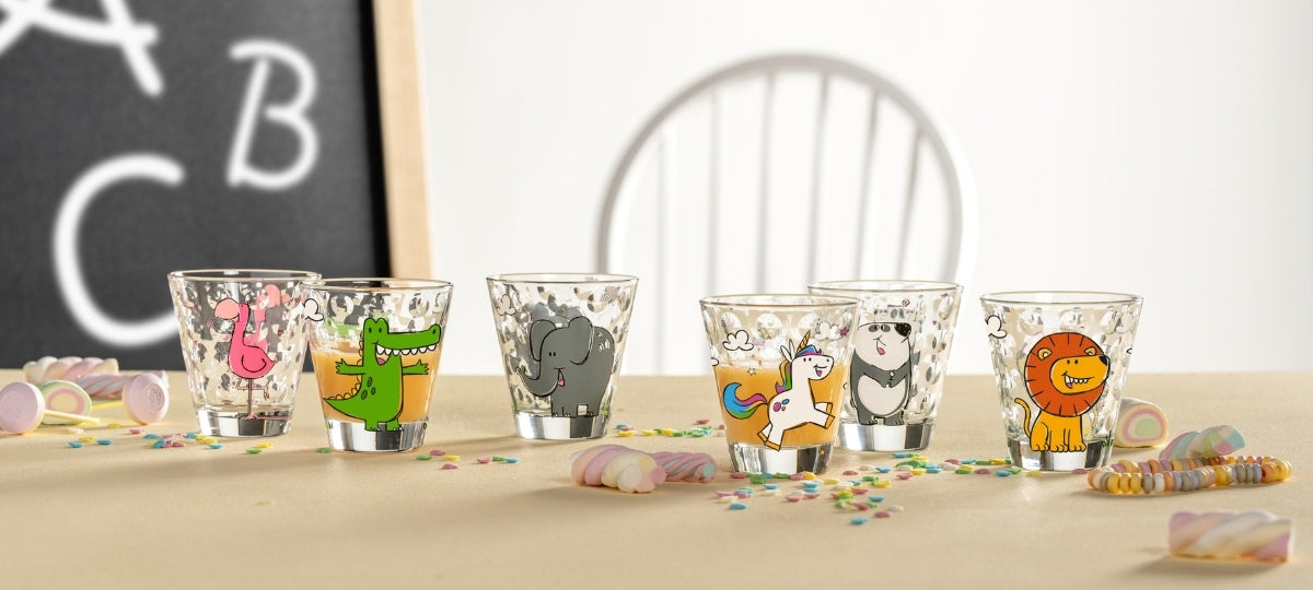 Bunte Kindergläser mit lustigen Tiermotiven wie Dinosaurier, Elefant, Löwe und Panda, umgeben von Konfetti und Perlenarmband auf beigem Untergrund
