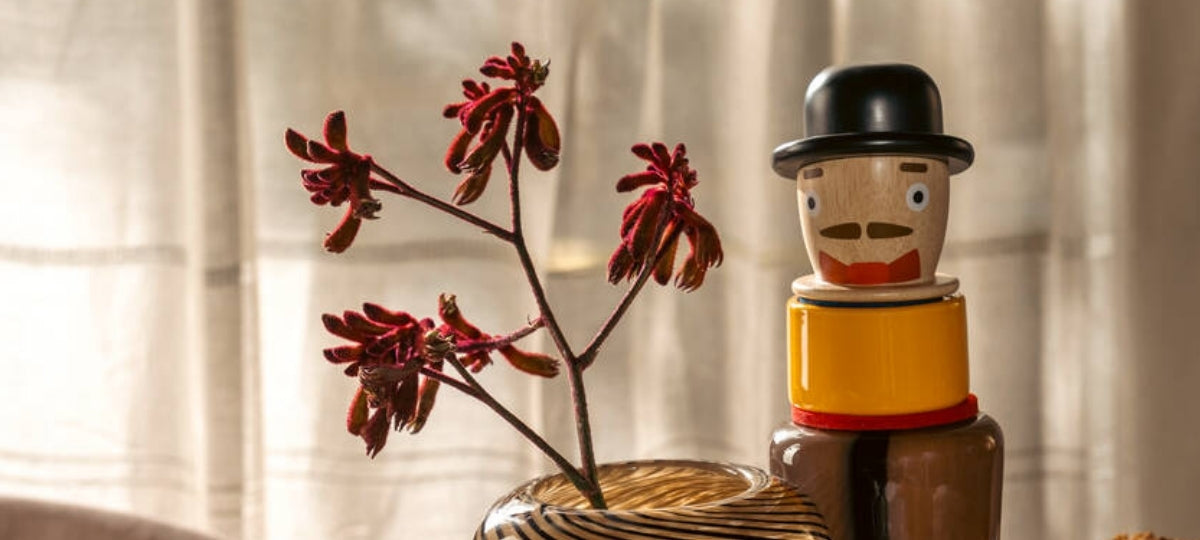 Holzfigur mit Hut neben getrockneter roter Blütenpflanze in Vase, helles Tageslicht, warme Farbtoene