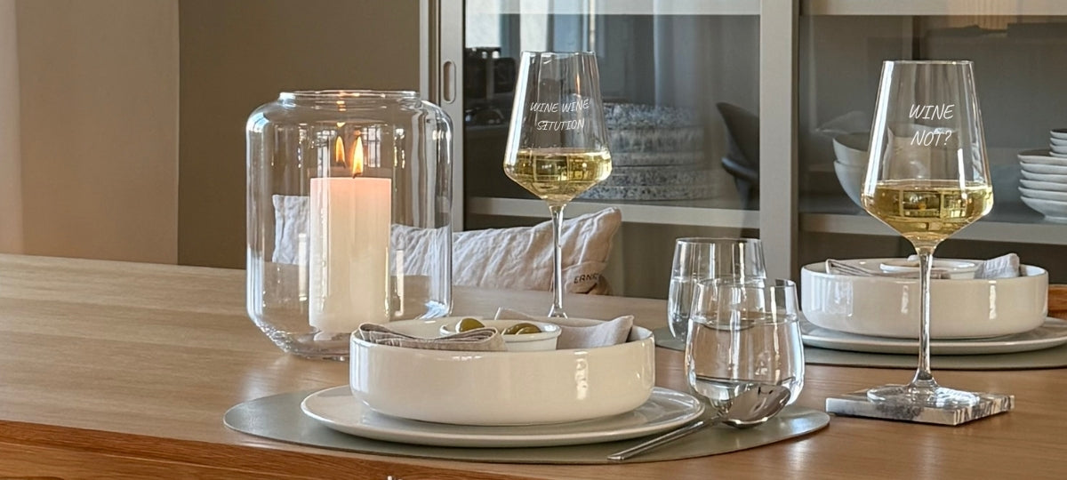 Elegante Tischdecke mit gravierten Weingläsern, Kerze in Glasvase, Tellern und cremefarbener Schale in warmem Licht