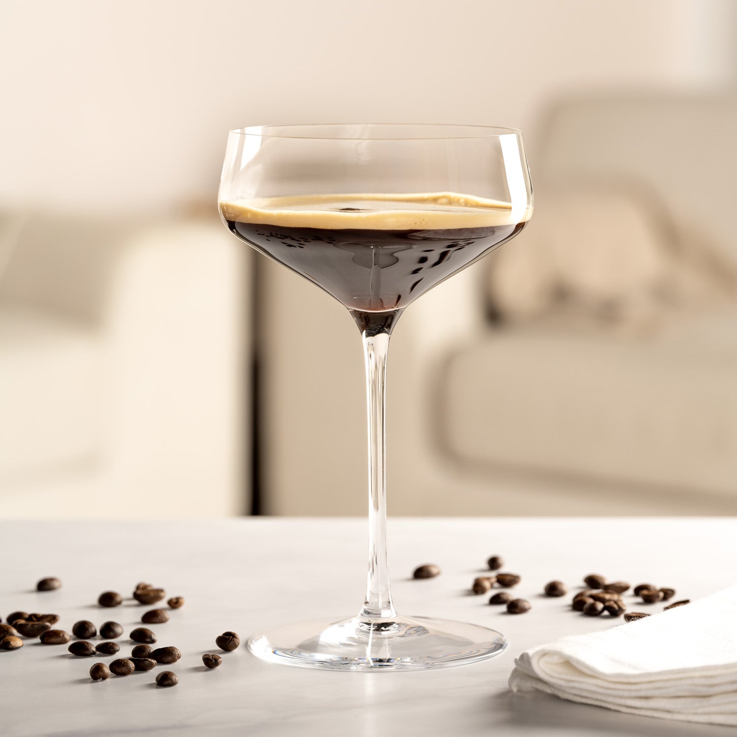 Klares Cocktailglas mit dunklem Espresso Martini auf einem Tisch, umgeben von Kaffeebohnen.