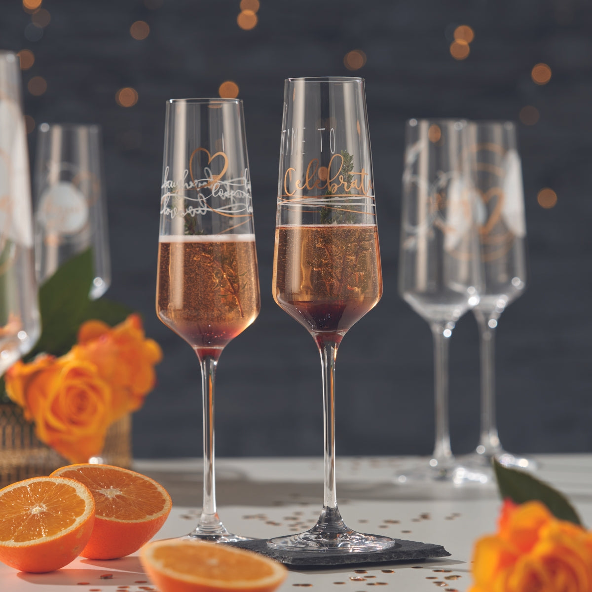 Zwei elegante Sektgläser mit goldener Schrift und prickelndem Drink, dekoriert mit Orangenscheiben und umgeben von leuchtenden Rosen und Konfetti.