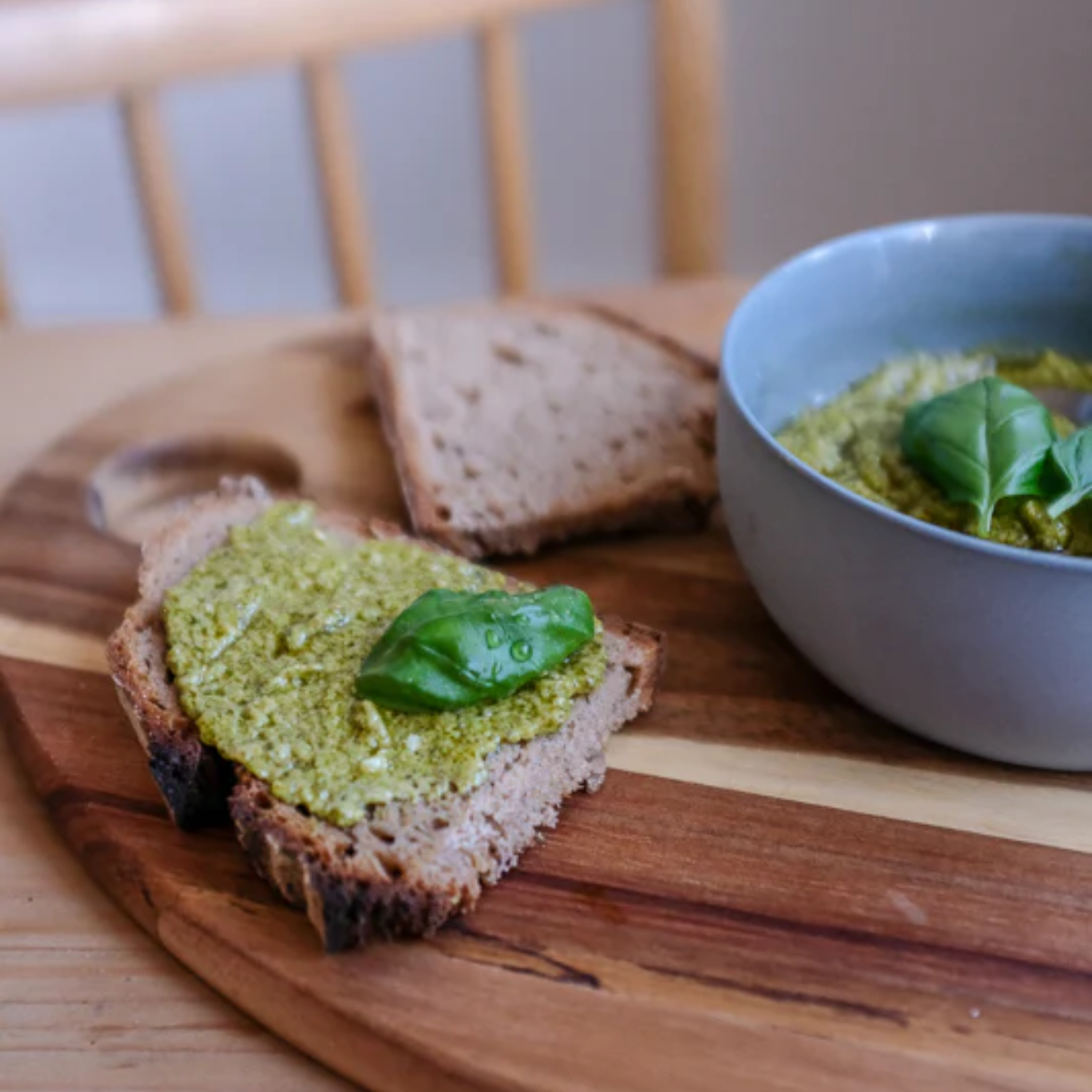 Eine Scheibe Vollkornbrot, dick mit grünem Pesto bestrichen und mit einem frischen Basilikumblatt garniert, liegt auf einem Holzbrett. Daneben eine graue Schale mit weiterem Pesto und eine zweite Brotscheibe im Hintergrund.