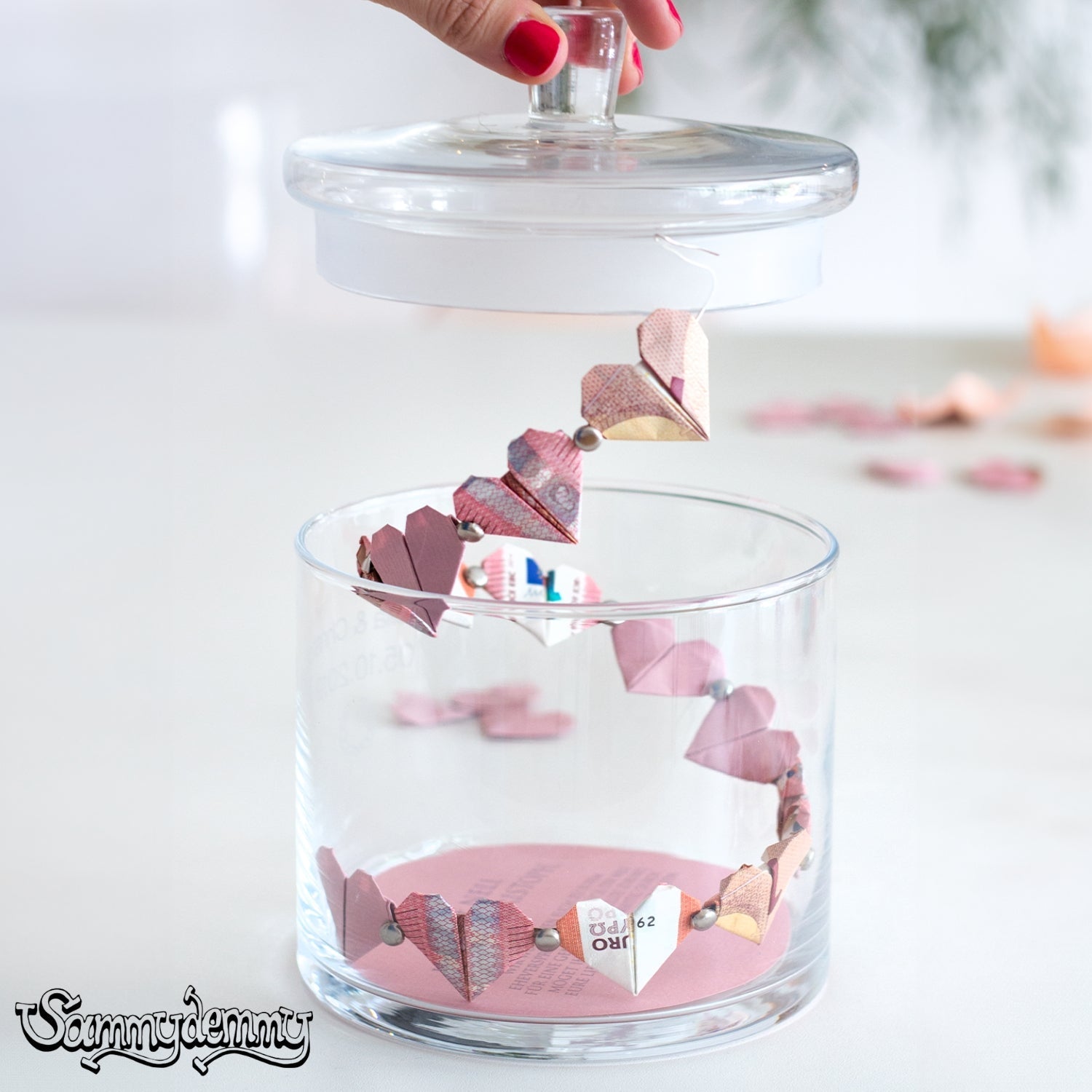Eine Glasdose mit einem Origami-Geldherz-Girlanden-Geschenk, bei dem die Herzchen von einem Draht an einem Deckel herunterhängen – eine kreative DIY-Idee für Hochzeiten oder besondere Anlässe.