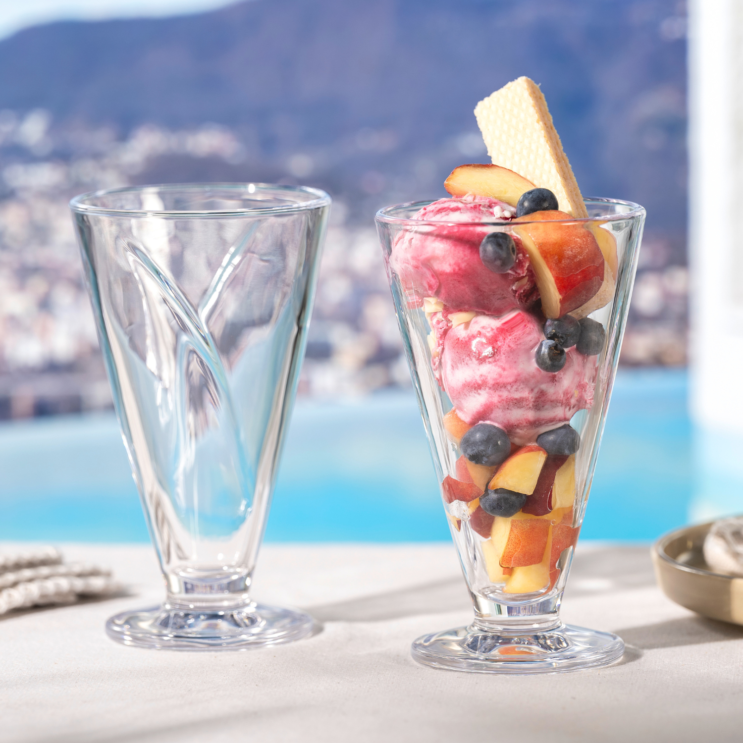 Zwei hohe Eisbecher stehen vor einer mediterranen Kulisse mit Poolblick – einer davon ist liebevoll mit Fruchteis, Blaubeeren, Nektarine und Waffel dekoriert.