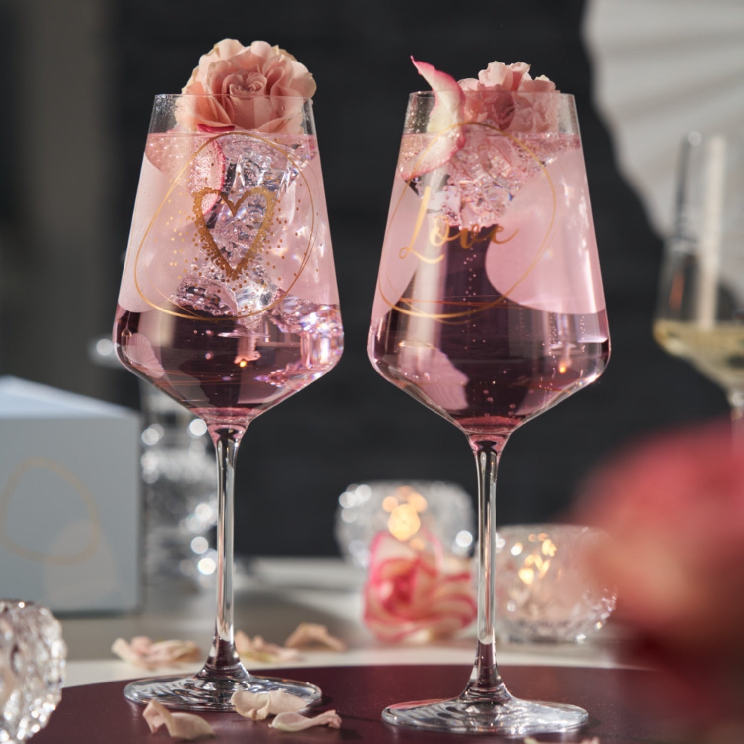 Zwei romantisch inszenierte Weingläser mit rosafarbenem Getränk, Eiswürfeln und rosa Blüten als Dekoration – perfekt für besondere Herzensmomente.