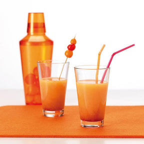 Trinkglas CIAO mit Orangensaft, Cocktailshaker und Strohhalmen auf orangefarbener Unterlage