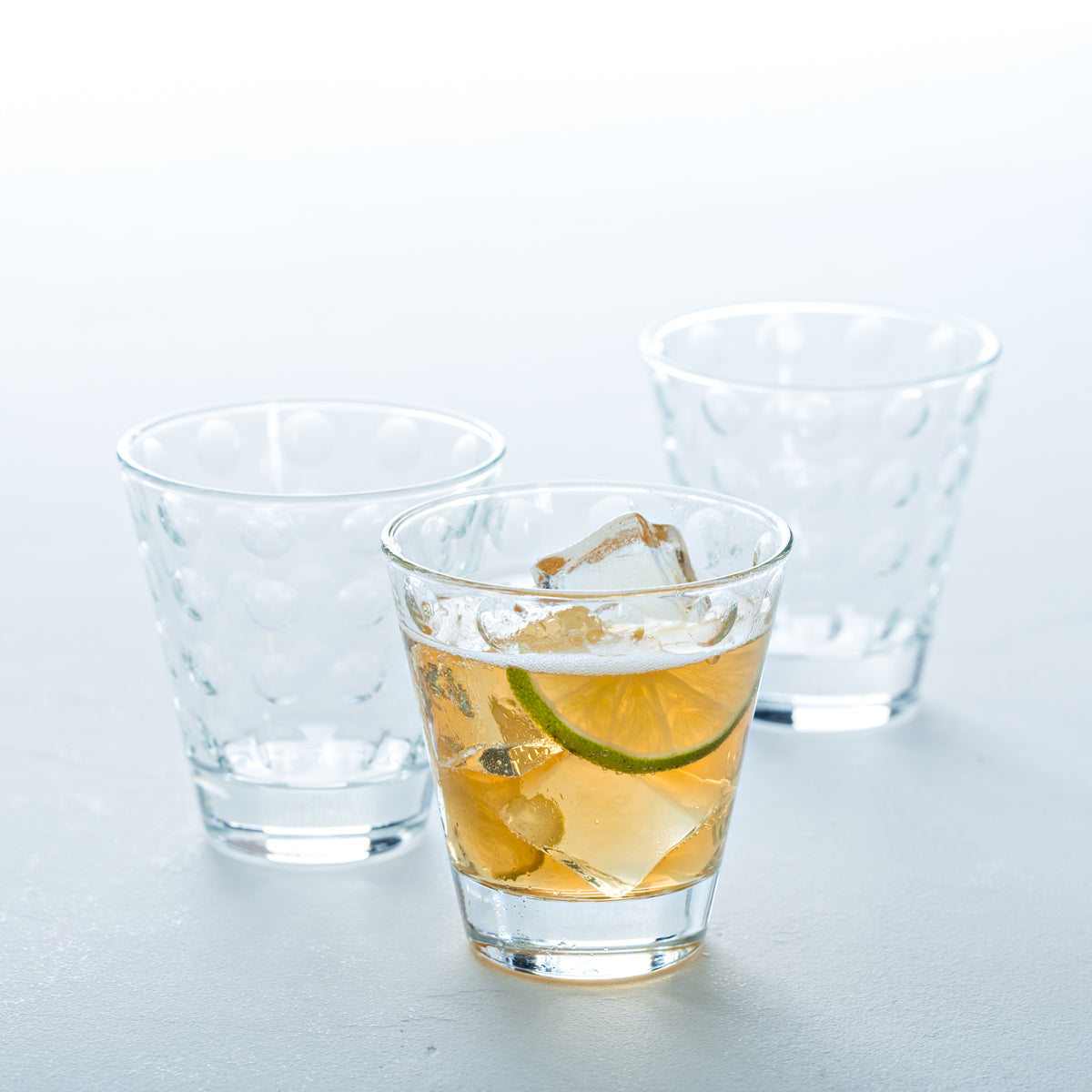 Drei Trinkgläser OPTIC, zwei leer und eines mit Getränk und Limettenscheibe gefüllt