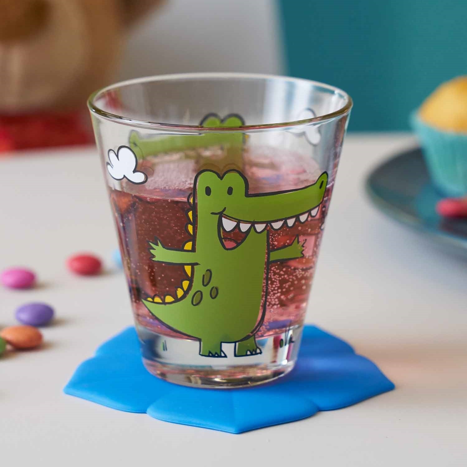 Trinkglas BAMBINI mit grünem Krokodil-Motiv, gefüllt mit Getränk auf blauem Untersetzer, bunte Perlen daneben