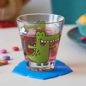 Trinkglas BAMBINI mit grünem Krokodil-Motiv, gefüllt mit Getränk auf blauem Untersetzer, bunte Perlen daneben