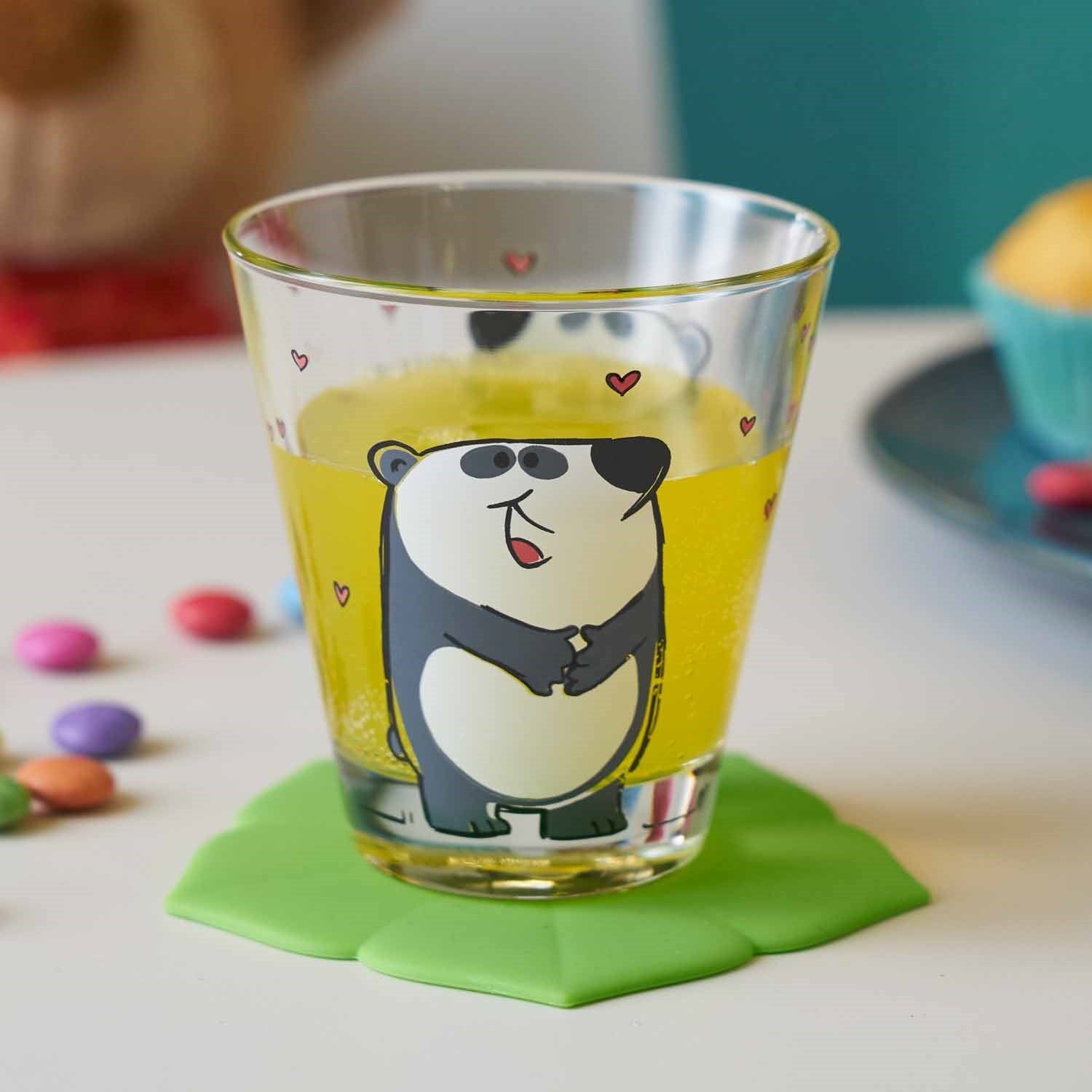 Trinkglas BAMBINI mit Panda-Motiv, gelbem Inhalt, auf grünem Untersetzer, umgeben von bunten Perlen