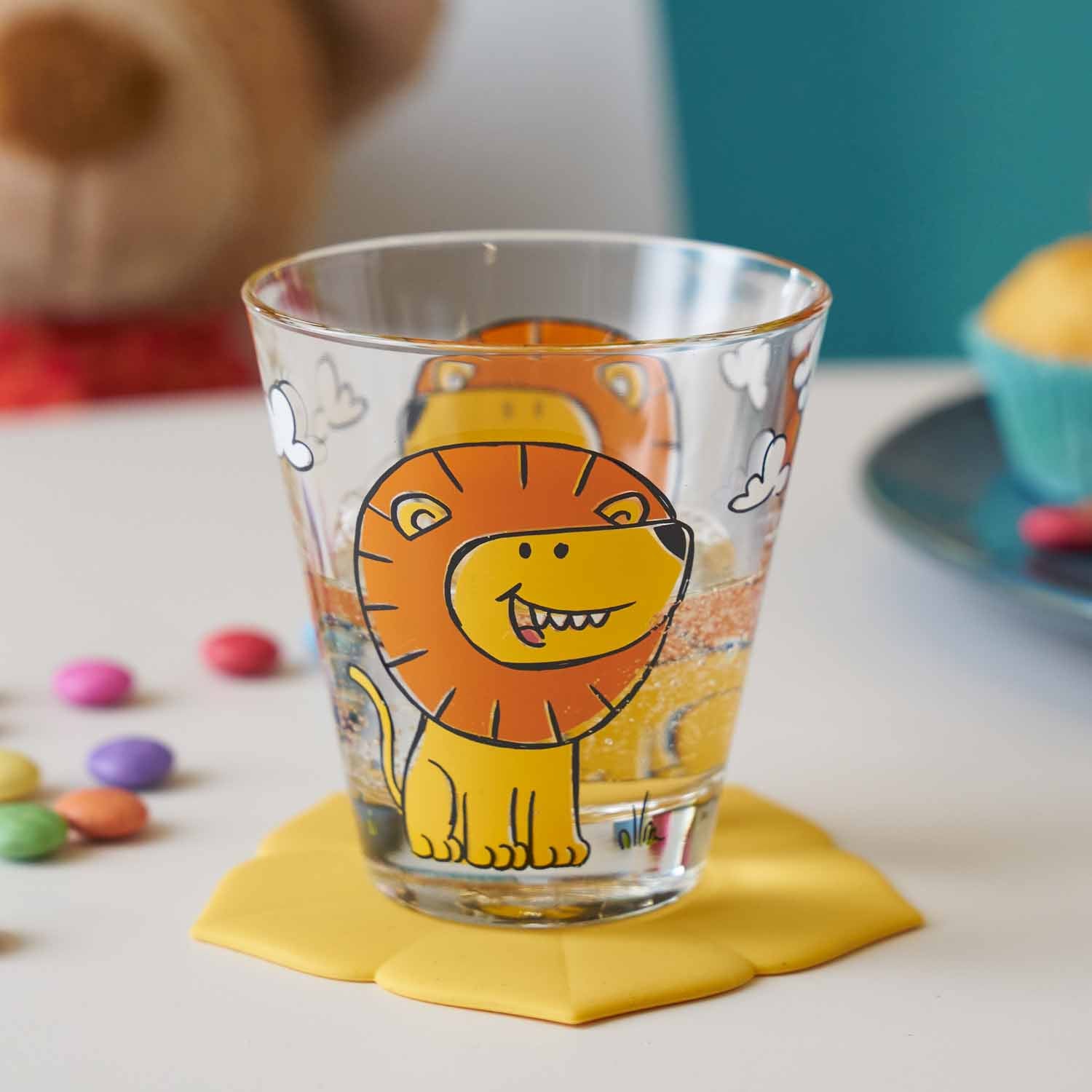 Kinderbecher BAMBINI mit Löwenmotiv auf gelbem Untersetzer, Bonbons und Spielzeug im Hintergrund