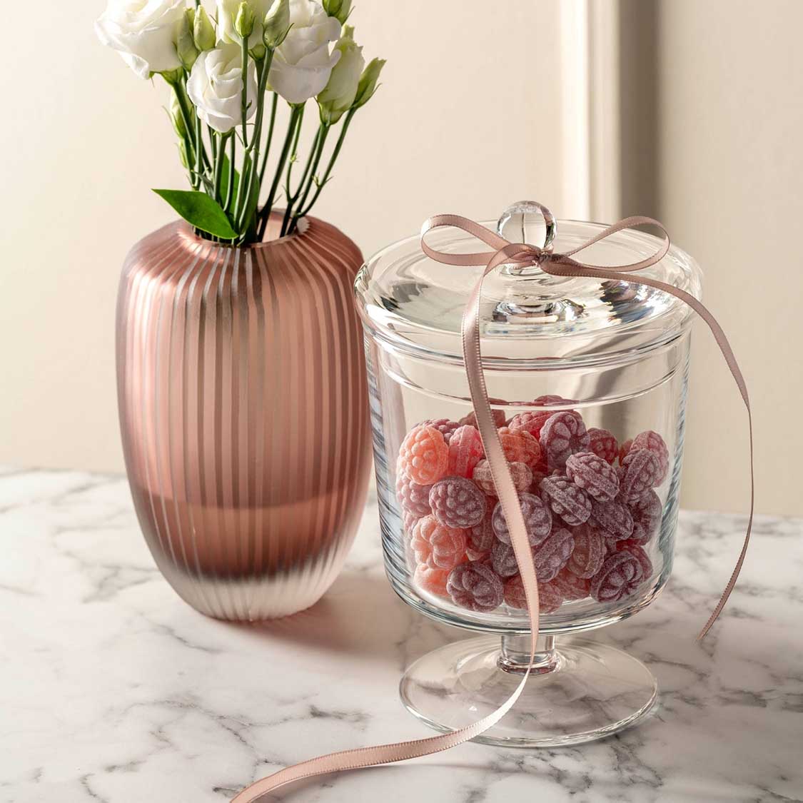 Glaskose POESIA mit Himbeeren, neben rosa Vase mit weißen Blüten auf Marmortisch