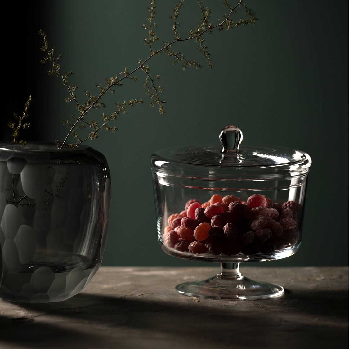Dose auf Fuß POESIA mit roten Trauben gefüllt, daneben schwarze Vase mit Zweigen, grüner Hintergrund