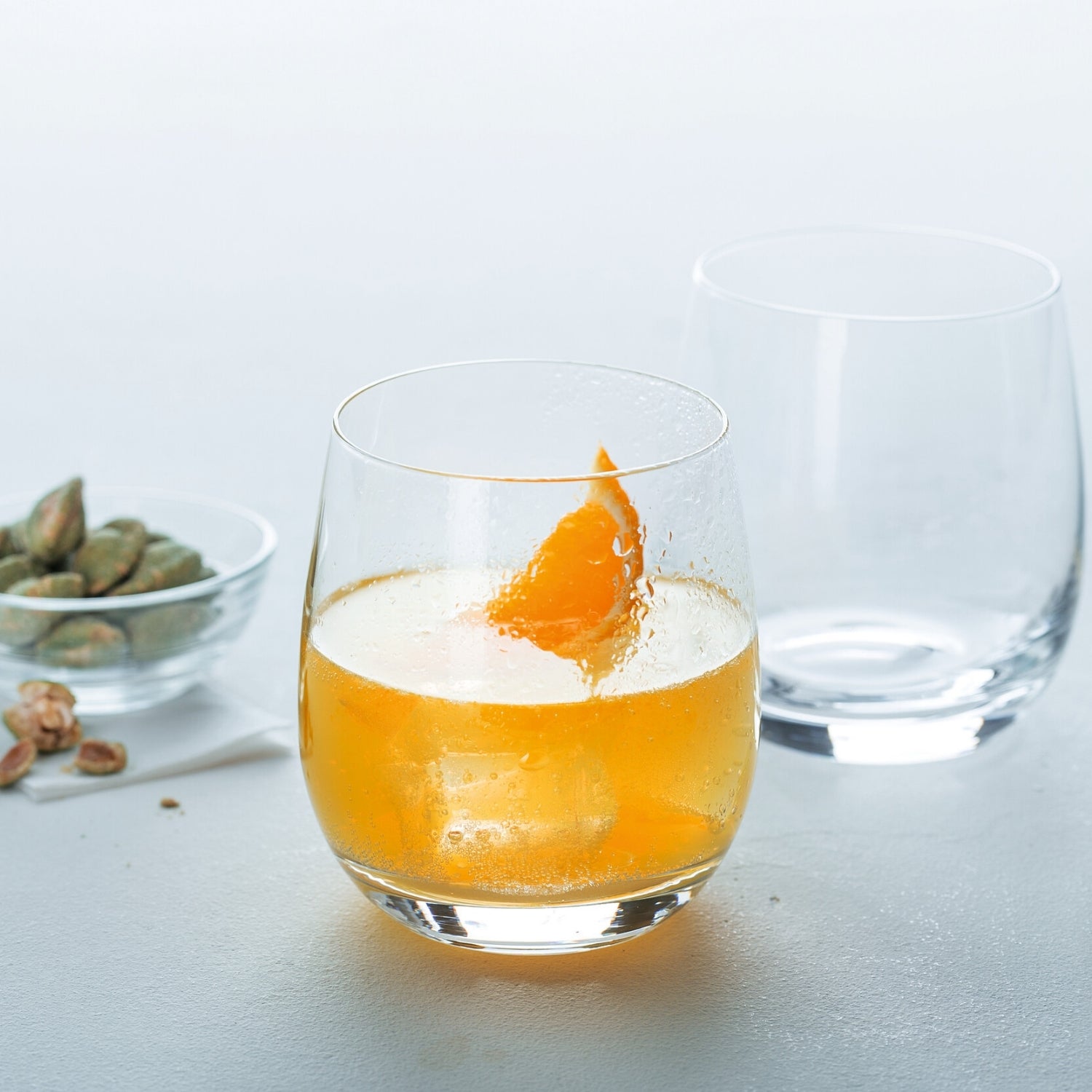 Trinkglas TIVOLI mit Orangensaft und Eiswürfeln, daneben leeres Glas und Schale mit Nüssen