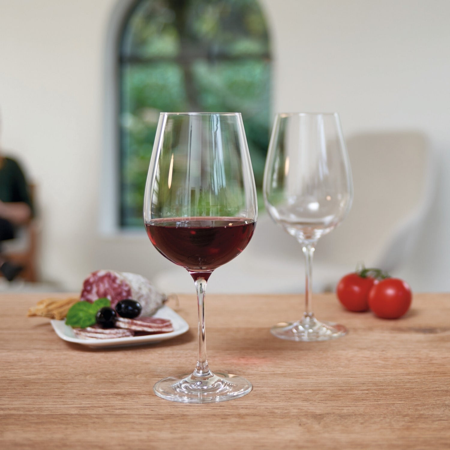 Rotweinglas TIVOLI mit Rotwein gefüllt auf Holztisch, Gedeck mit Wurstplatte und Tomaten im Hintergrund