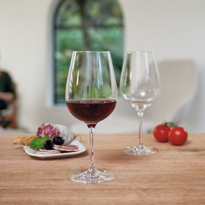 Rotweinglas TIVOLI mit Rotwein gefüllt auf Holztisch, Gedeck mit Wurstplatte und Tomaten im Hintergrund
