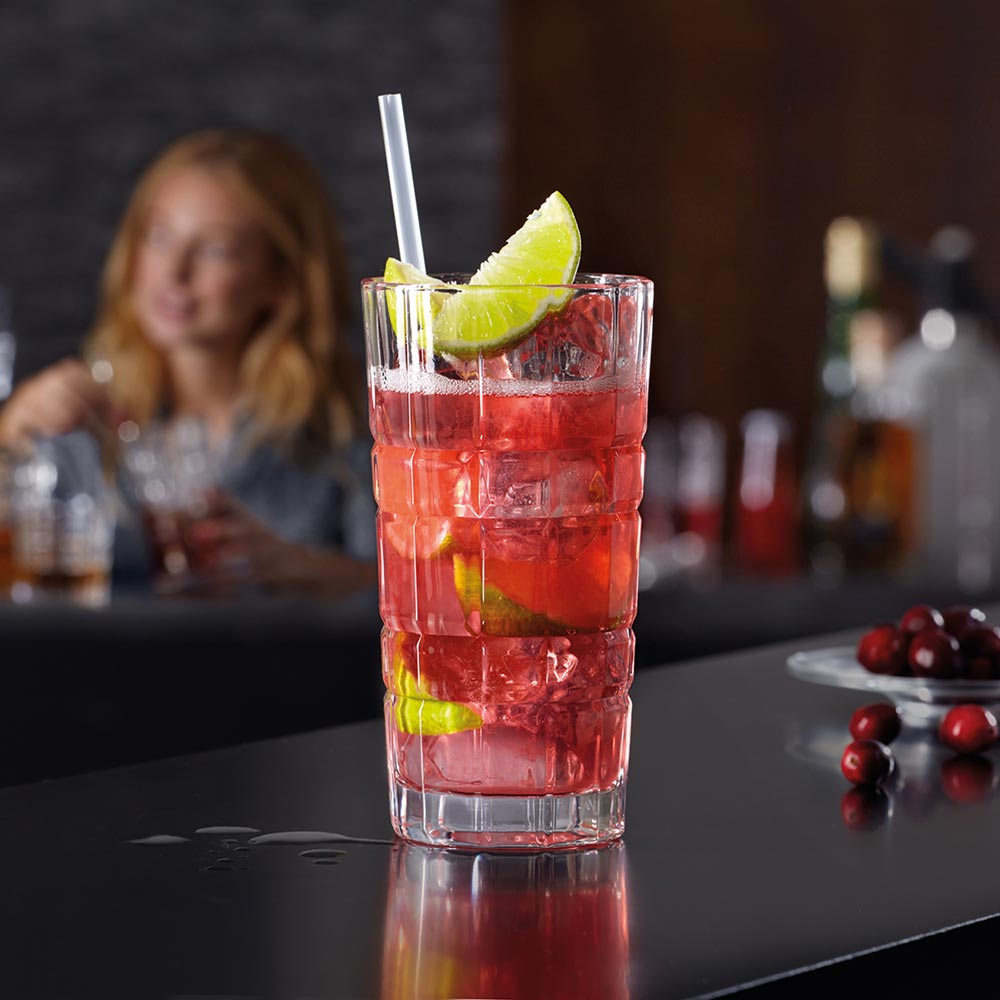 Longdrinkglas SPIRITII mit rotem Cocktail, Limettenscheibe und Strohhalm, Barszene im Hintergrund