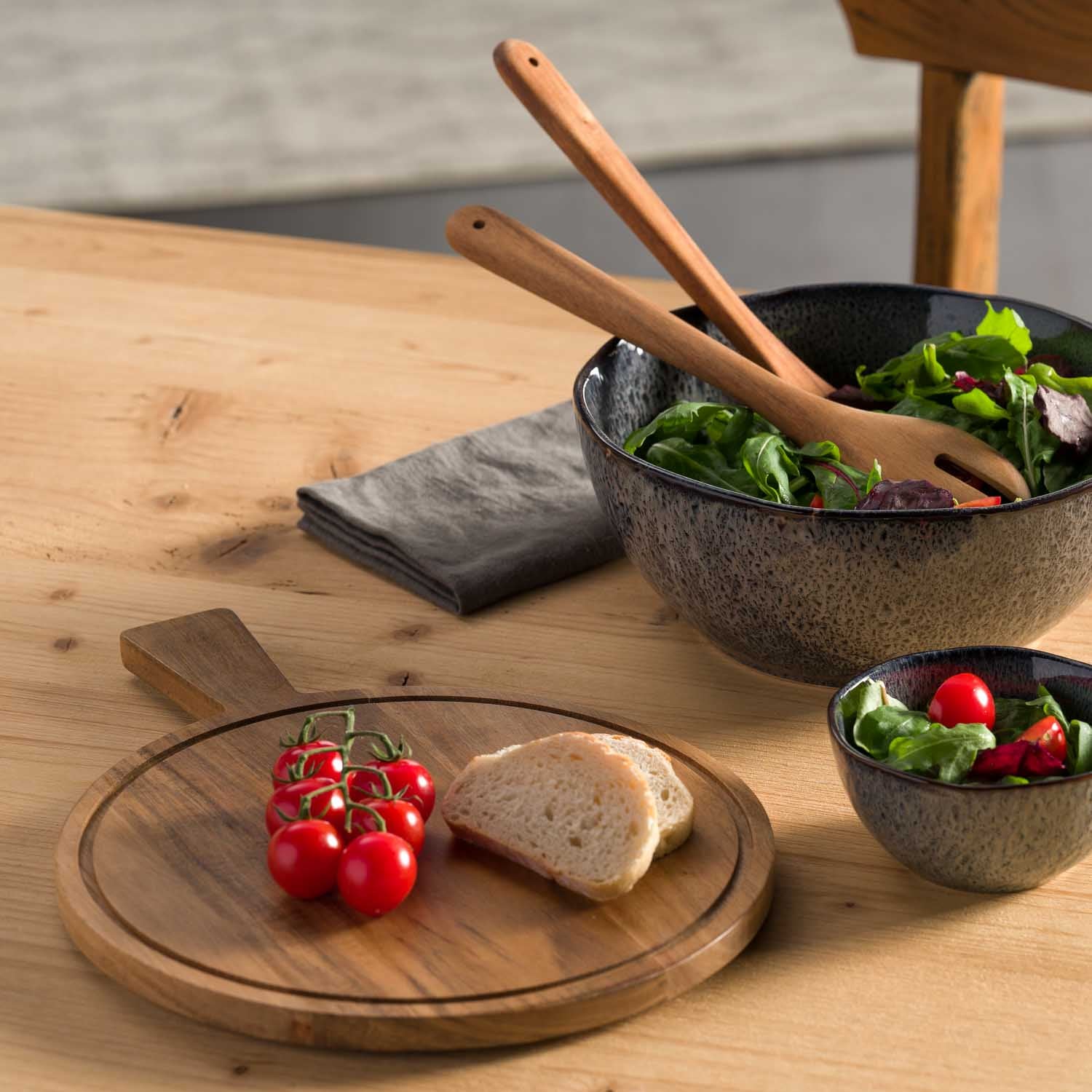 Schneidebrett MATERA mit Tomaten und Brot, Schüsseln mit Salat und Kräutern auf Holztisch