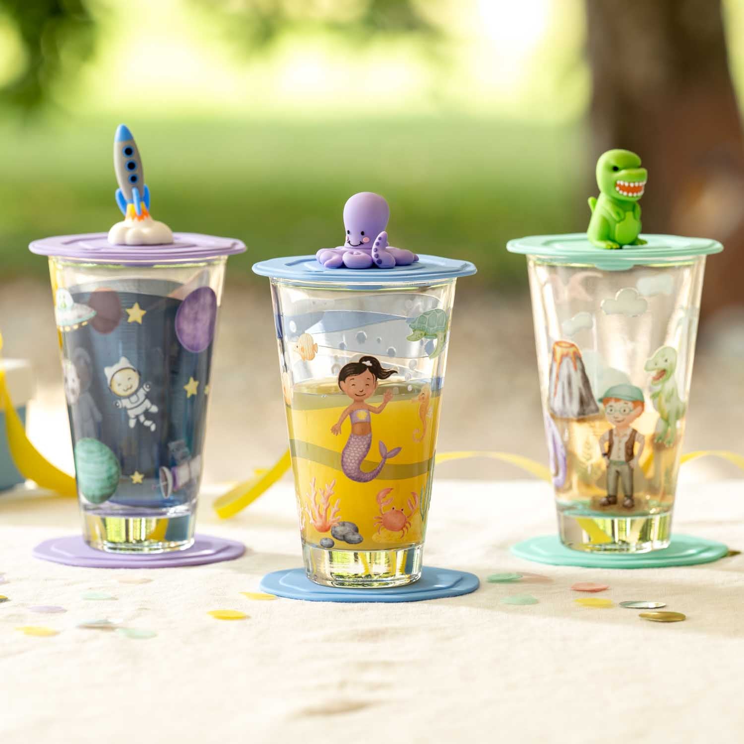 Drei Glasbecher mit bunten Motiven und Deckeln im Freien auf Tisch