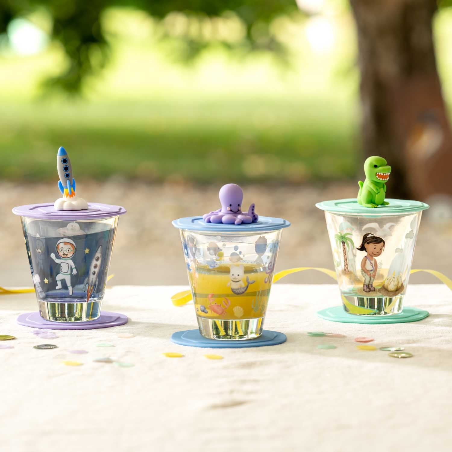 Drei bunte Trinkgläser mit Dino-Motiven und Figuren-Deckeln im Freien auf Tisch