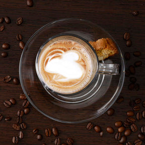 Glastasse SENSO mit Latte-Macchiato, umgeben von Kaffeebohnen und Gebäck