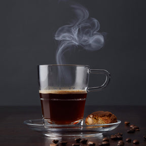 Glastasse mit heißem Kaffee und Untertasse SENSO, Kaffeebohnen und Gebäck daneben