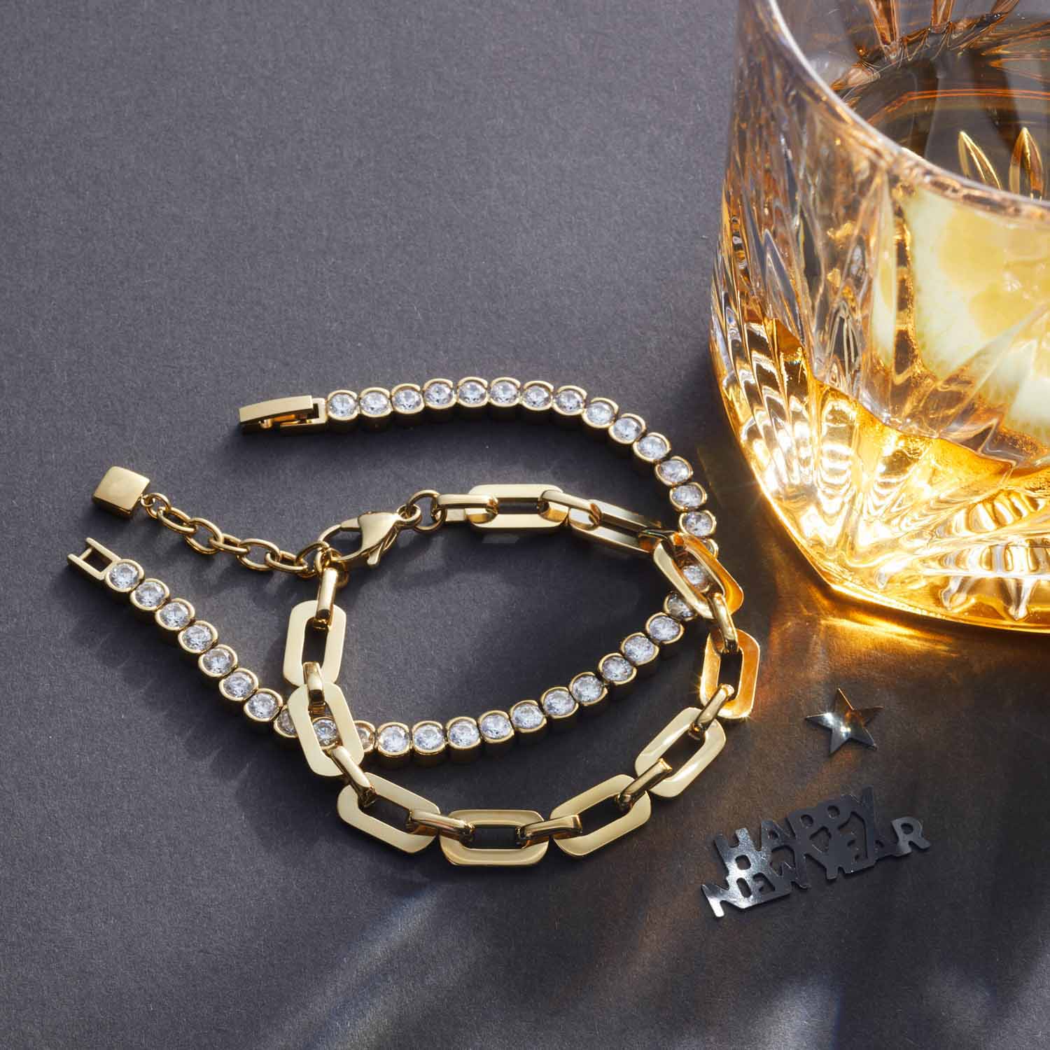Armband CREMONA gold mit Glaskristallen und Kettengliedern neben Whiskeyglas auf grauem Untergrund