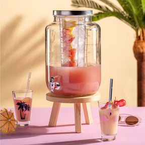 Getränkespender SUCCO mit rosa Getränk, gefüllt mit Früchten, auf rosa Tisch mit Gläsern