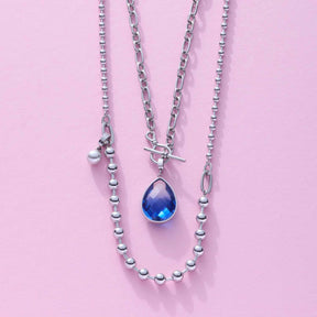 Halskette ELISABETH mit silbernen Kugelketten und blauem Tropfenanhänger auf rosa Hintergrund