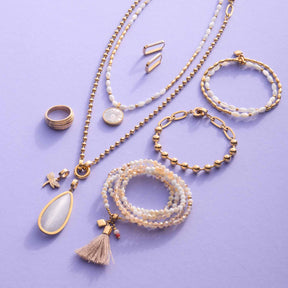 Schmuckkollektion mit Halsketten, Armbändern, Ring und Ohrsteckern in Gold und Perltönen auf lila Hintergrund