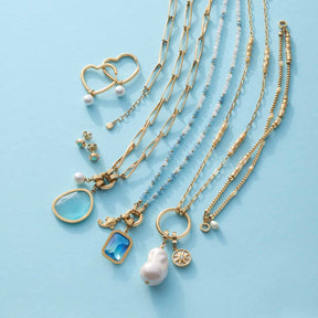 Creole LULU gold mit verschiedenen Halsketten und Schmuckzubehoer auf hellblauem Untergrund