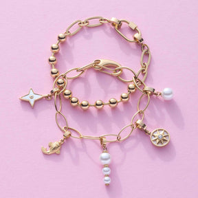 Drei goldene Armbänder der Serie TORY mit Perlen und Charms auf rosa Hintergrund