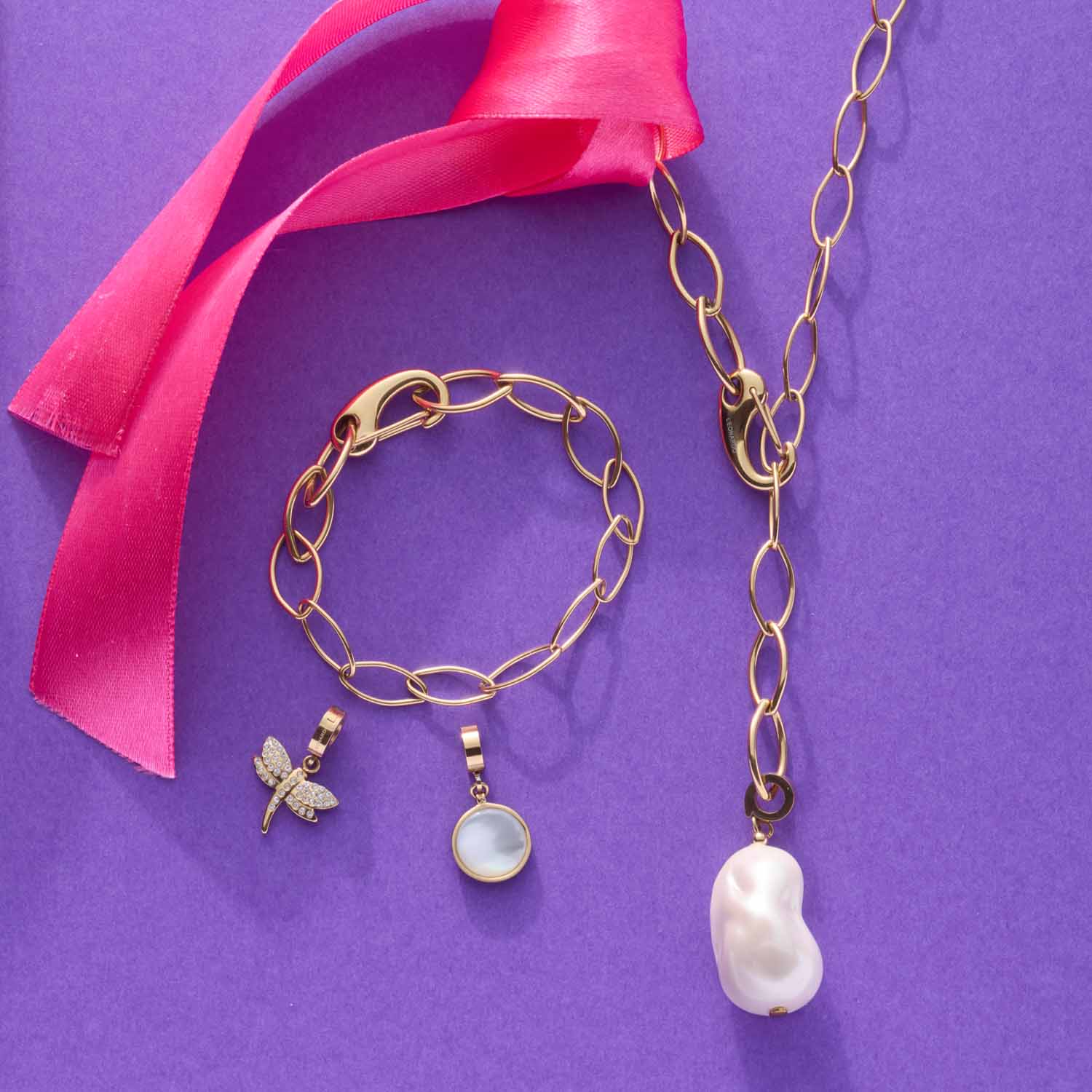 Armband TORY gold mit Kettengliedern, passenden Anhängern und rosa Band auf violettem Hintergrund