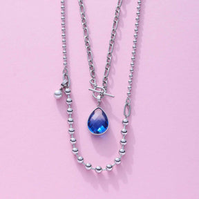 Halskette DOTTY mit silbernen Kugelgliedern und blauem Tropfenanhänger auf rosa Hintergrund