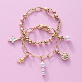 Drei goldene Armbänder DOTTY mit Perlen und Charms auf rosa Hintergrund