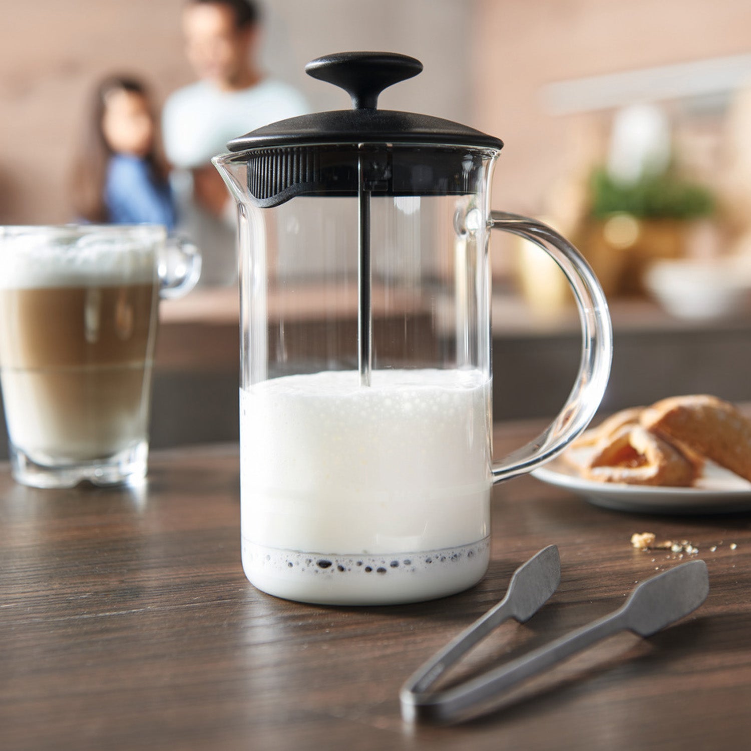 Milchaufschäumer CAFFEE PER ME mit aufgeschäumter Milch auf Küchentheke, Glas daneben, Gebäck und Personen im Hintergrund