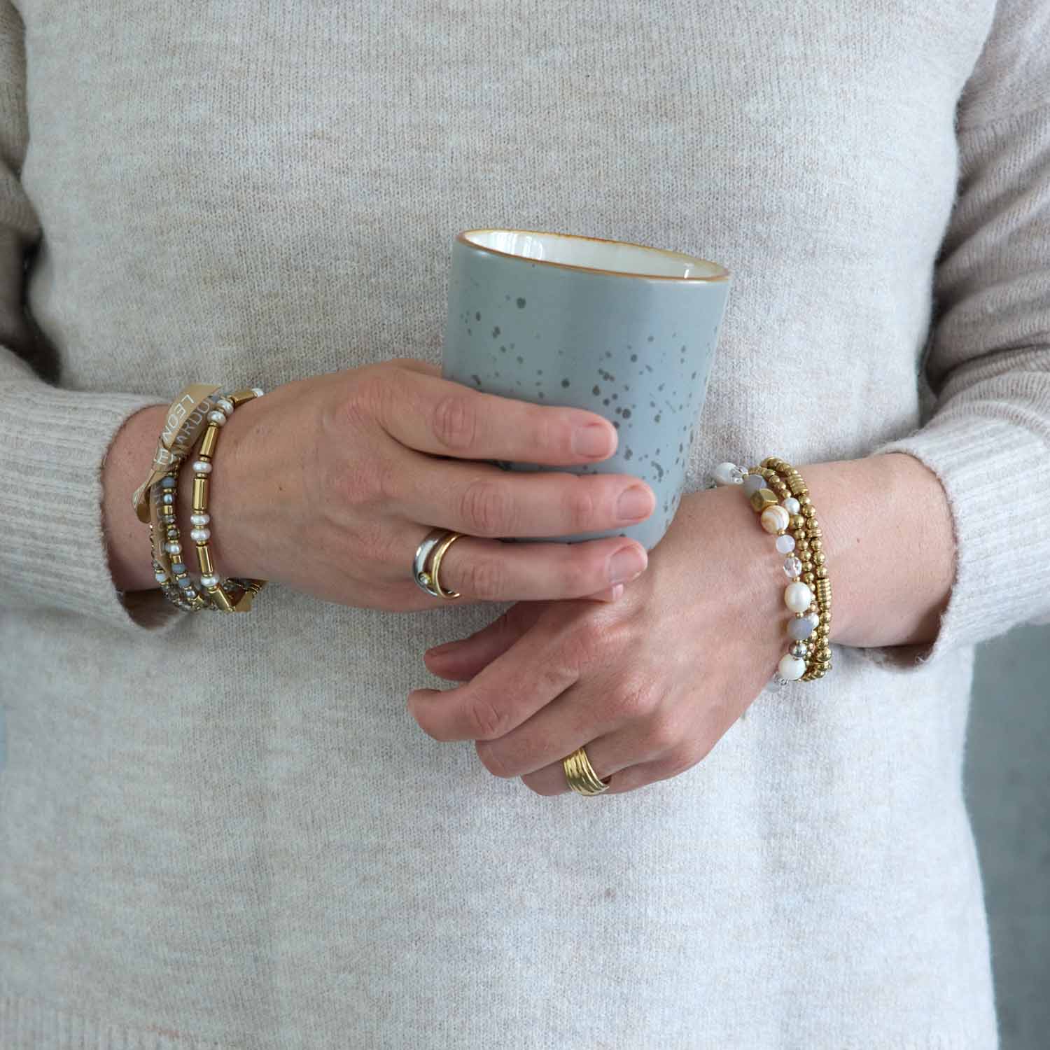 Frau traegt mehrere gold-farbene Armbaender BERNIE Clip & Mix an beiden Handgelenken, haelt hellblaue Tasse