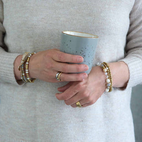 Frau traegt mehrere gold-farbene Armbaender BERNIE Clip & Mix an beiden Handgelenken, haelt hellblaue Tasse