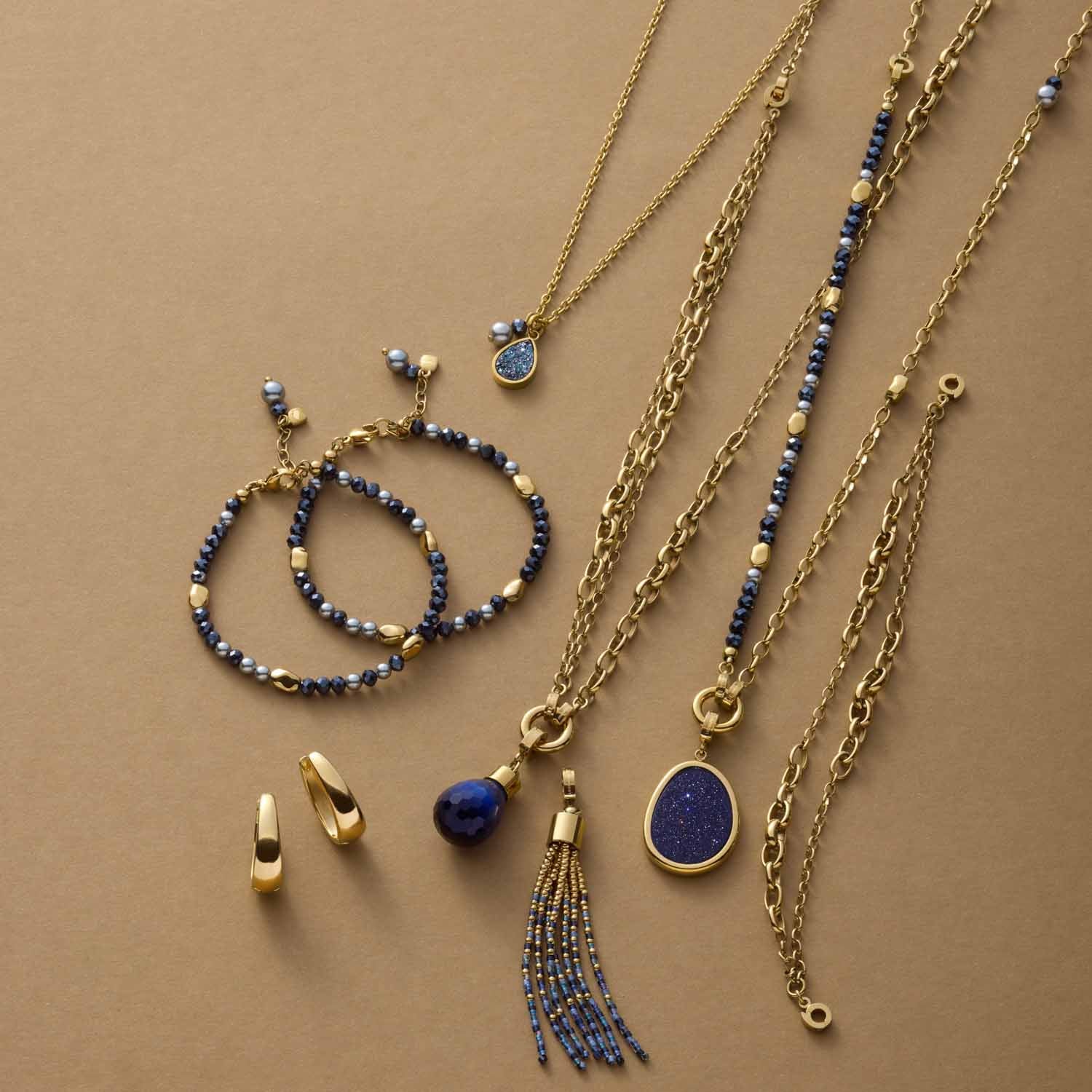 Goldene Schmuckkollektion mit Armbändern, Ketten, Ohrringen und blauen Steinen auf beigem Hintergrund