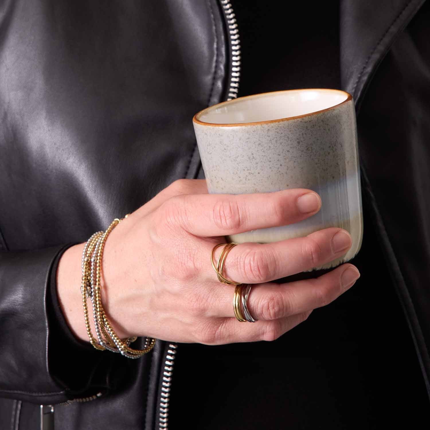 Person in schwarzer Jacke hält beigefarbenes Glas, traegt Armband NOEMI gold CIAO
