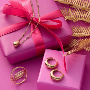 Halskette CLOTHILDE mit Anhänger auf rosa Geschenkbox neben Ringen und goldenem Blattdekor