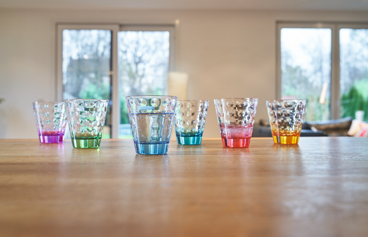 Sechs Trinkgläser OPTIC 215 ml in verschiedenen Farben auf Holztisch, Fenster im Hintergrund