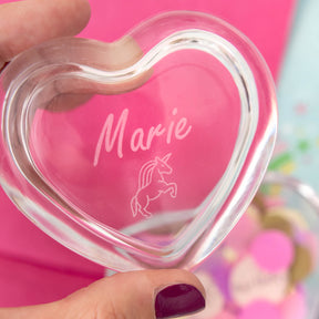 Herzdose HEART in Glasform mit Gravur "Marie" und Einhorn-Symbol, in Hand gehalten vor rosa Hintergrund