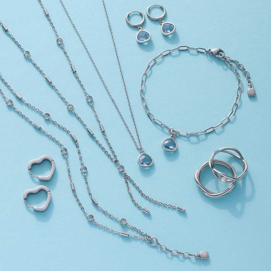 Silberfarbene Schmuckkollektion mit Halsketten, Ringen, Armbändern und Ohrringen auf hellblauem Hintergrund