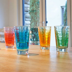 Vier bunte Trinkgläser mit verschiedenfarbigen Getränken auf Holztisch, Fenster im Hintergrund