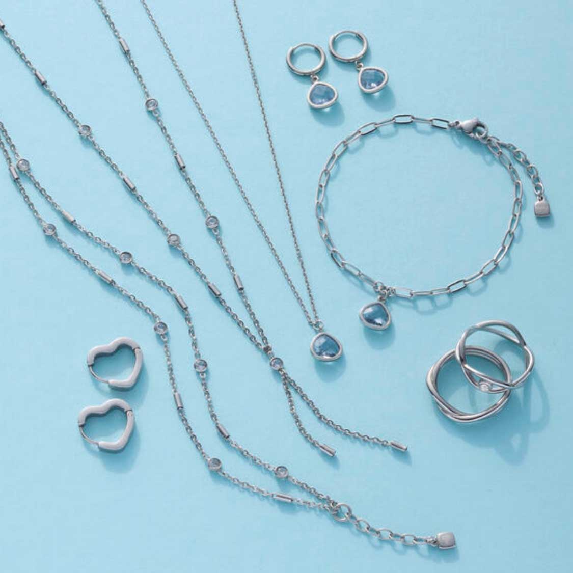 Silberner Schmuck aus der Serie LUVELLA: Creolen, Halsketten, Ringe und Kettenarmband auf hellblauem Untergrund