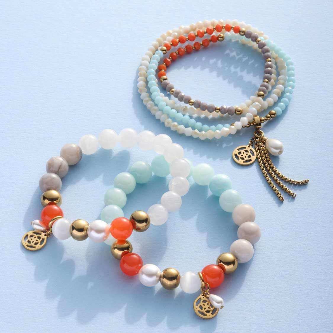 Mehrere Glasperlen-Armbänder in Blau-, Weiß- und Orangetönen mit goldenen Metallperlen und Anhängern auf hellem Hintergrund